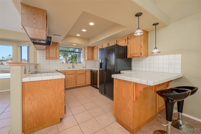 kitchen featuring tile counters, a raised ceiling, hanging light fixtures, a peninsula, and black appliances