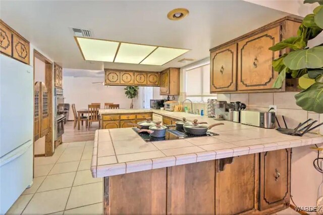 kitchen with a peninsula, tile counters, brown cabinets, and freestanding refrigerator