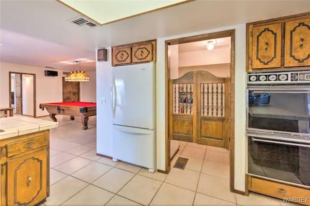 kitchen featuring tile countertops, light tile patterned floors, freestanding refrigerator, multiple ovens, and decorative light fixtures