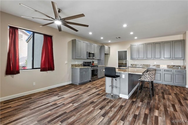 kitchen featuring light stone counters, gray cabinetry, appliances with stainless steel finishes, an island with sink, and a kitchen breakfast bar