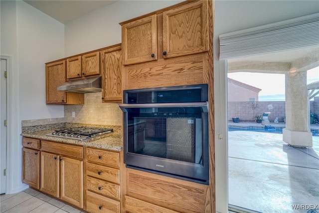 kitchen featuring stainless steel appliances, backsplash, brown cabinetry, and under cabinet range hood