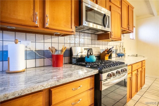 kitchen with light tile patterned floors, light stone counters, appliances with stainless steel finishes, decorative backsplash, and brown cabinetry