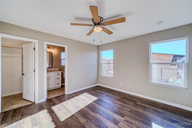 unfurnished bedroom featuring ensuite bathroom, a ceiling fan, baseboards, a spacious closet, and dark wood-style floors