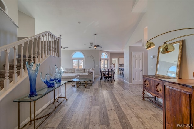 unfurnished living room with baseboards, light wood-style flooring, ceiling fan, stairs, and high vaulted ceiling