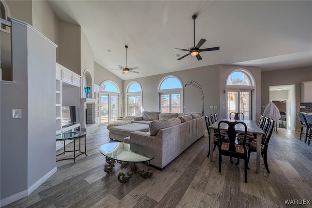 living room featuring baseboards, a fireplace with raised hearth, dark wood-style floors, ceiling fan, and high vaulted ceiling