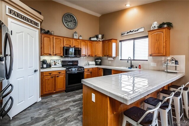 kitchen with tile countertops, a peninsula, a sink, brown cabinets, and black appliances