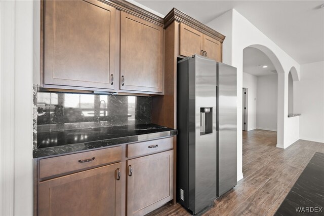 kitchen featuring tasteful backsplash, arched walkways, stainless steel fridge with ice dispenser, dark wood-type flooring, and dark stone countertops