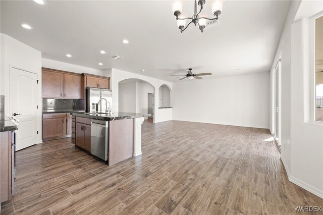 kitchen with arched walkways, dark wood finished floors, an island with sink, open floor plan, and stainless steel appliances