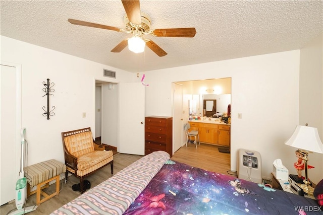 bedroom with a textured ceiling, ceiling fan, ensuite bathroom, wood finished floors, and visible vents