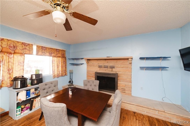 dining space with a textured ceiling, a stone fireplace, wood finished floors, and a ceiling fan