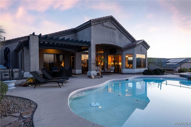 pool at dusk with a patio, grilling area, fence, exterior kitchen, and an outdoor pool