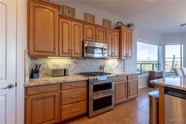 kitchen with light tile patterned floors, appliances with stainless steel finishes, backsplash, light stone countertops, and brown cabinetry