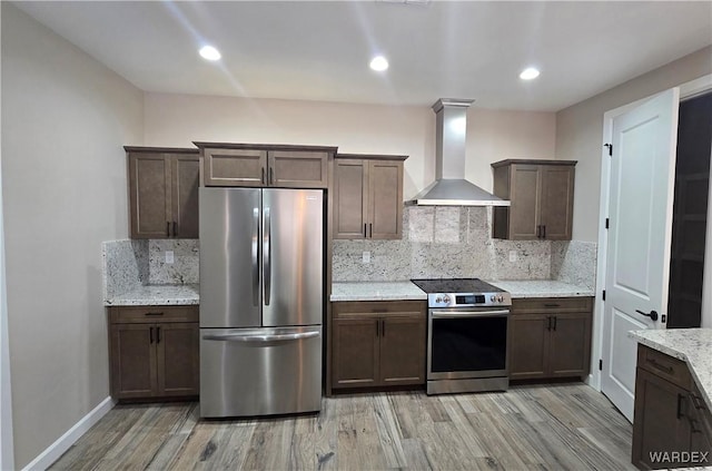 kitchen with light wood finished floors, wall chimney exhaust hood, appliances with stainless steel finishes, light stone counters, and backsplash