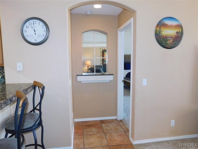 hallway featuring arched walkways, light colored carpet, baseboards, and light tile patterned floors