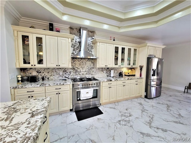 kitchen with appliances with stainless steel finishes, glass insert cabinets, wall chimney exhaust hood, and light stone countertops