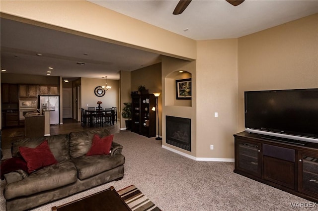 living room with ceiling fan with notable chandelier, dark colored carpet, a glass covered fireplace, and baseboards