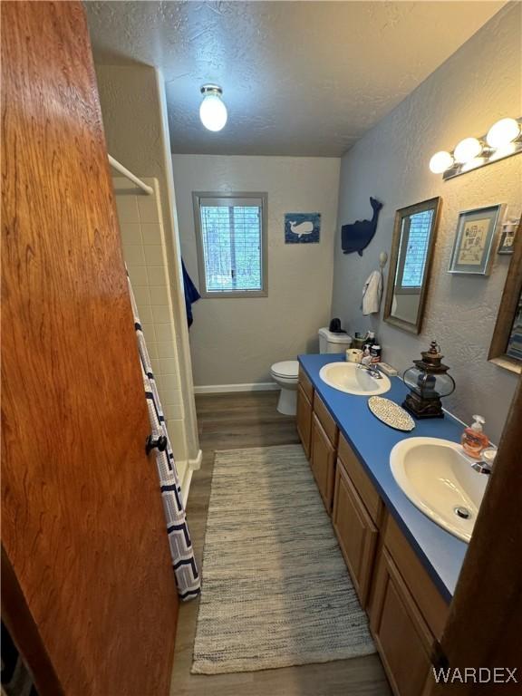 full bath with a textured ceiling, a textured wall, toilet, a sink, and double vanity