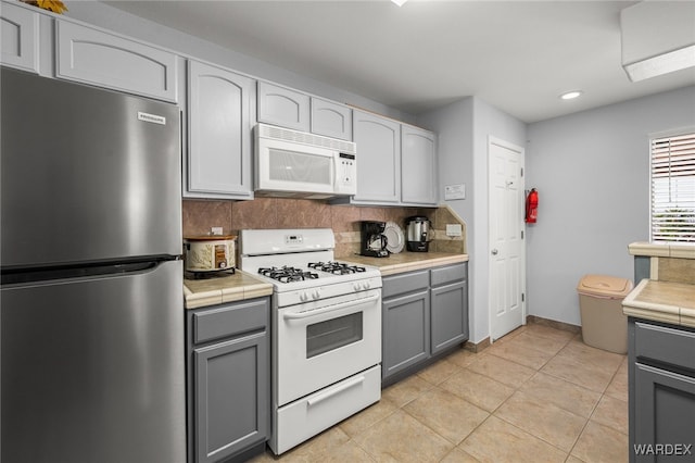 kitchen featuring tile countertops, light tile patterned floors, gray cabinets, backsplash, and white appliances