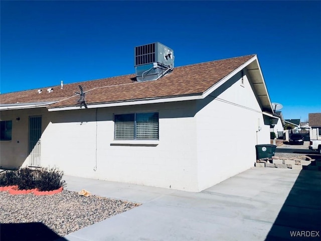 view of side of home with concrete block siding, central AC, a patio, and roof with shingles