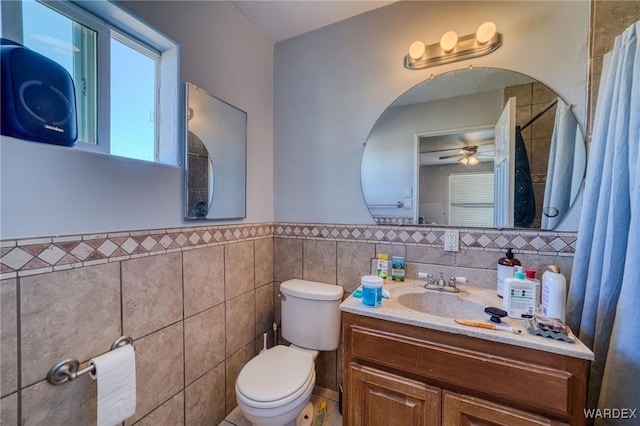 bathroom featuring a ceiling fan, toilet, a wainscoted wall, vanity, and tile walls