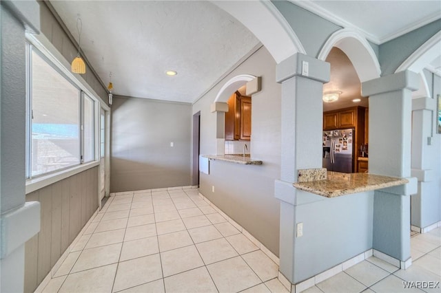 kitchen with stainless steel fridge, arched walkways, brown cabinetry, light stone counters, and light tile patterned flooring