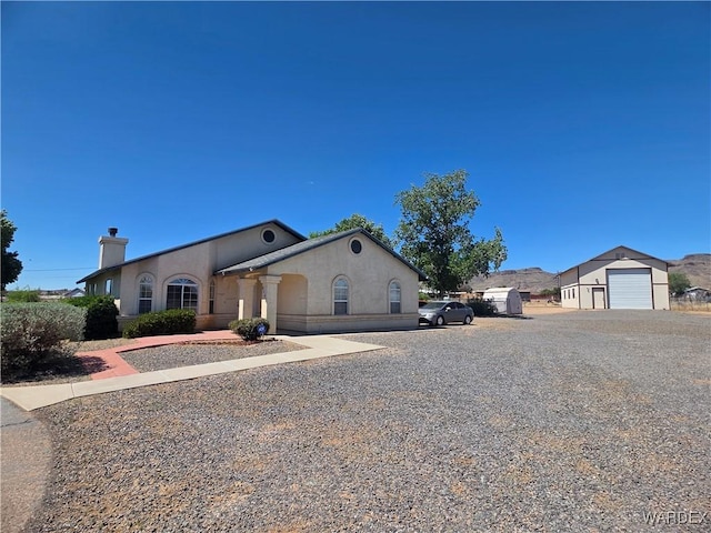 single story home featuring a detached garage, gravel driveway, a chimney, and stucco siding