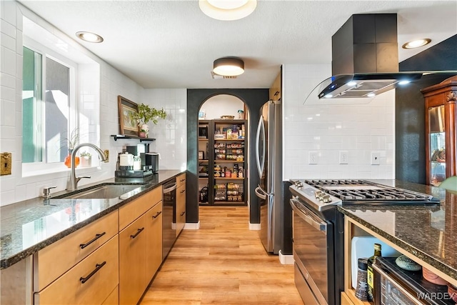 kitchen with appliances with stainless steel finishes, a sink, island range hood, dark stone counters, and dark cabinetry