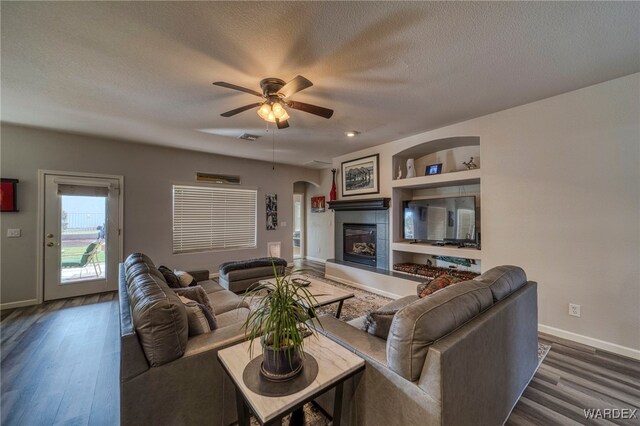 living room with built in shelves, a glass covered fireplace, dark wood-style flooring, and a textured ceiling