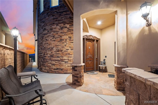 exterior entry at dusk with stone siding, visible vents, fence, and stucco siding