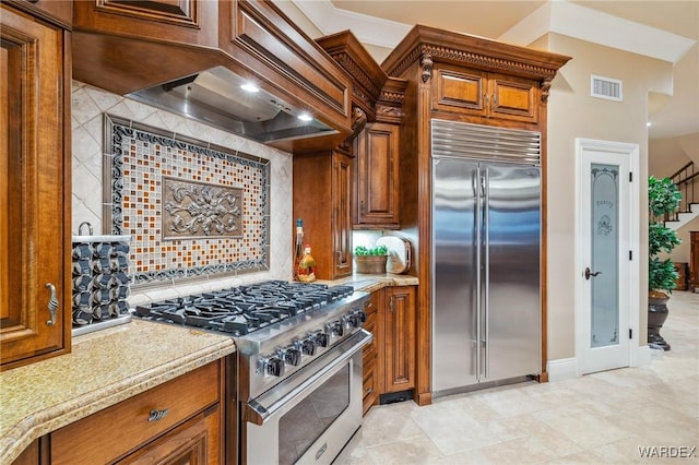 kitchen with high end appliances, custom range hood, visible vents, decorative backsplash, and brown cabinetry