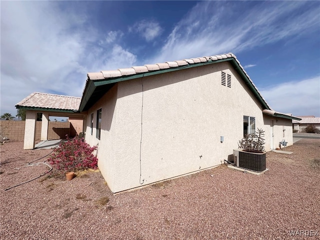 view of side of home with central AC, fence, a tiled roof, and stucco siding