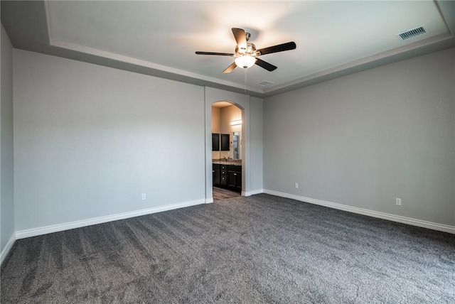 empty room with arched walkways, a raised ceiling, dark colored carpet, a ceiling fan, and baseboards