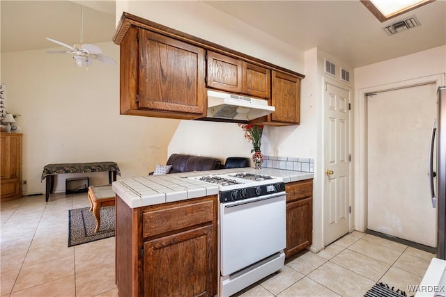 kitchen with brown cabinets, white gas stove, tile countertops, visible vents, and under cabinet range hood