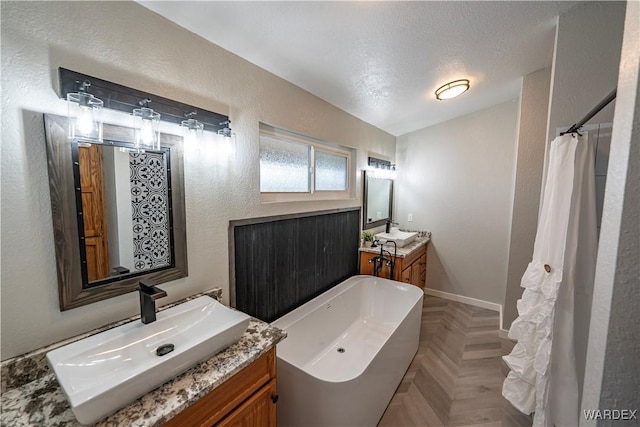 full bathroom with a textured ceiling, a textured wall, two vanities, a sink, and a soaking tub