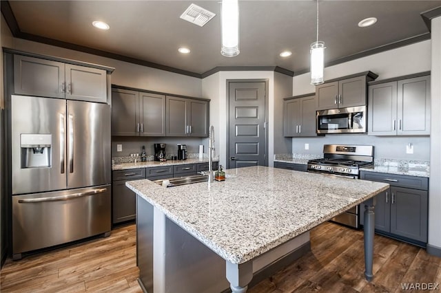 kitchen with a center island with sink, hanging light fixtures, appliances with stainless steel finishes, a sink, and light stone countertops