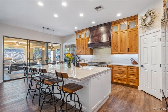 kitchen with glass insert cabinets, visible vents, a center island with sink, decorative light fixtures, and custom range hood