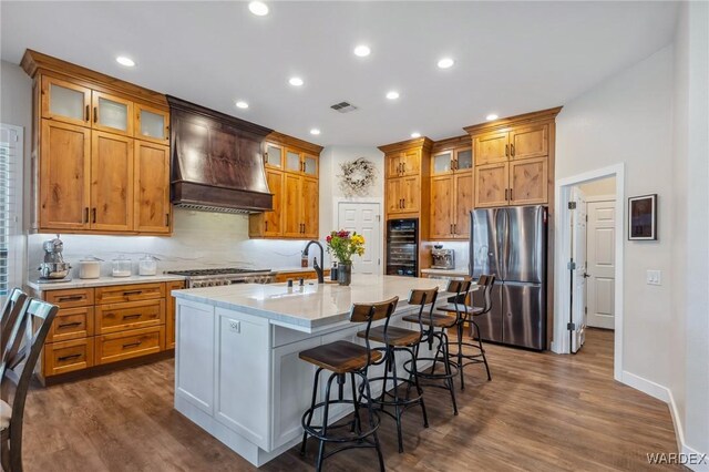 kitchen featuring stainless steel appliances, visible vents, glass insert cabinets, an island with sink, and premium range hood