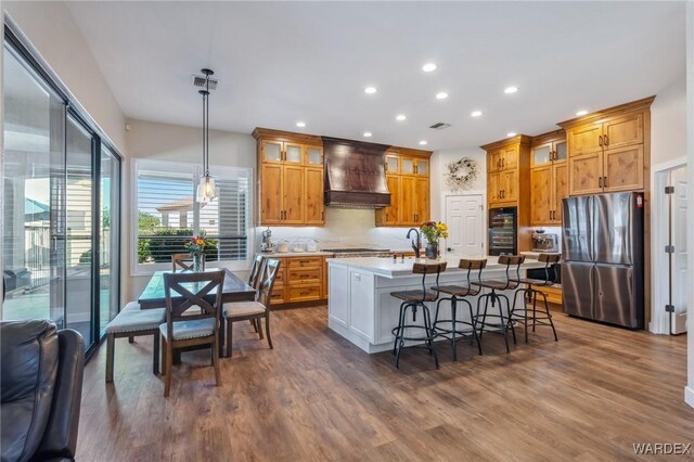 kitchen with appliances with stainless steel finishes, hanging light fixtures, a kitchen island with sink, light countertops, and premium range hood