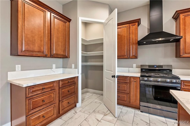 kitchen with stainless steel range with gas cooktop, marble finish floor, brown cabinetry, light stone countertops, and wall chimney exhaust hood