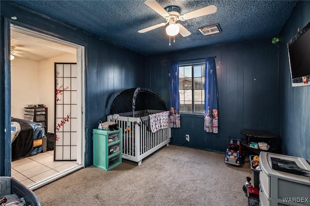 carpeted bedroom with a textured ceiling, ceiling fan, a nursery area, and visible vents