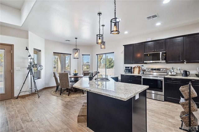 kitchen with an island with sink, appliances with stainless steel finishes, decorative light fixtures, wood finish floors, and a sink