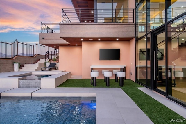 back of house at dusk featuring a patio, a balcony, an in ground hot tub, an outdoor pool, and stucco siding