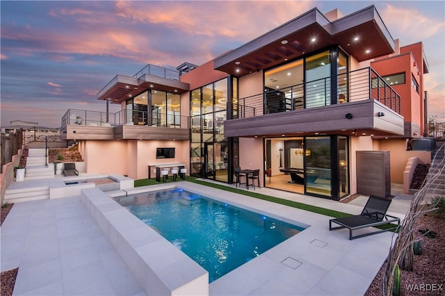 rear view of house with an infinity pool, a patio, stucco siding, a hot tub, and a balcony