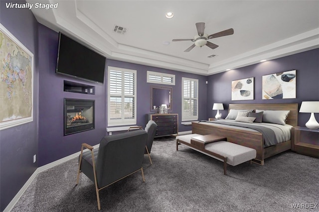 bedroom featuring a raised ceiling, visible vents, dark carpet, a glass covered fireplace, and baseboards