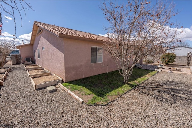 view of side of home featuring stucco siding, a tile roof, central AC, fence, and a vegetable garden