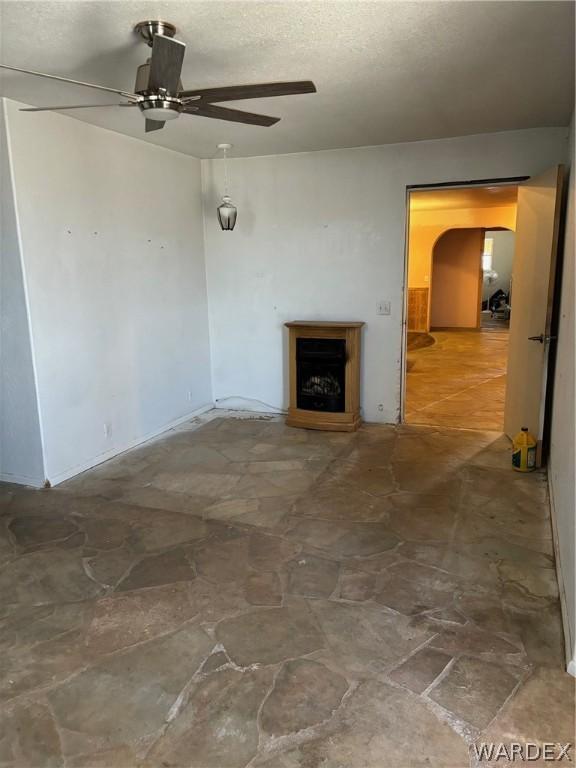 unfurnished living room with a fireplace with raised hearth, stone finish floor, a textured ceiling, and a ceiling fan