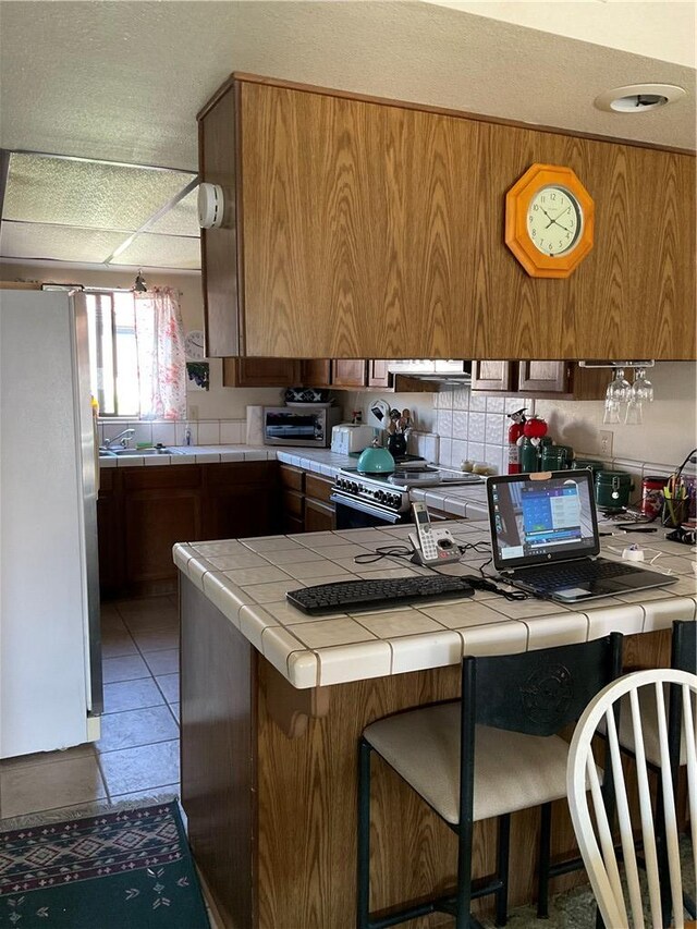201 Riverfront Drive, Drive, Bullhead City, AZ 86442 - kitchen with freestanding refrigerator, tile countertops, brown cabinetry, high end stainless steel range, and a peninsula
