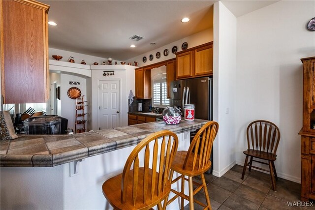 kitchen featuring a peninsula, visible vents, freestanding refrigerator, tile counters, and brown cabinetry