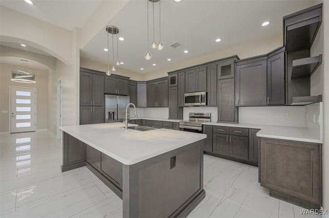 kitchen with arched walkways, stainless steel appliances, light countertops, an island with sink, and pendant lighting