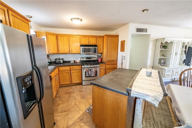 kitchen featuring stainless steel appliances, dark countertops, visible vents, brown cabinetry, and a textured ceiling